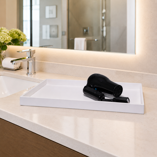 White | A white rectangular tray on a beige bathroom countertop holding a black hair dryer and a small black accessory, with a sink, faucet, mirror, and flowers in the background.
